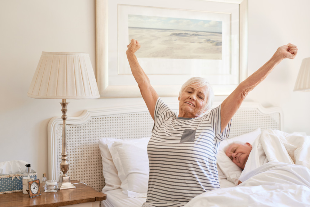 senior woman stretching on the edge of her bed in the morning with husband sleeping
