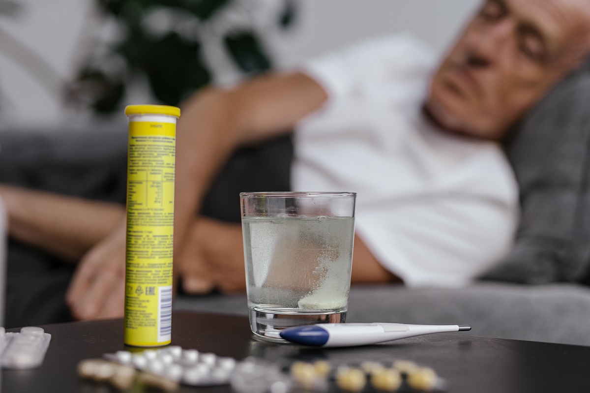 medicines and a glass of water on a table, with an older man lying on a couch in the background